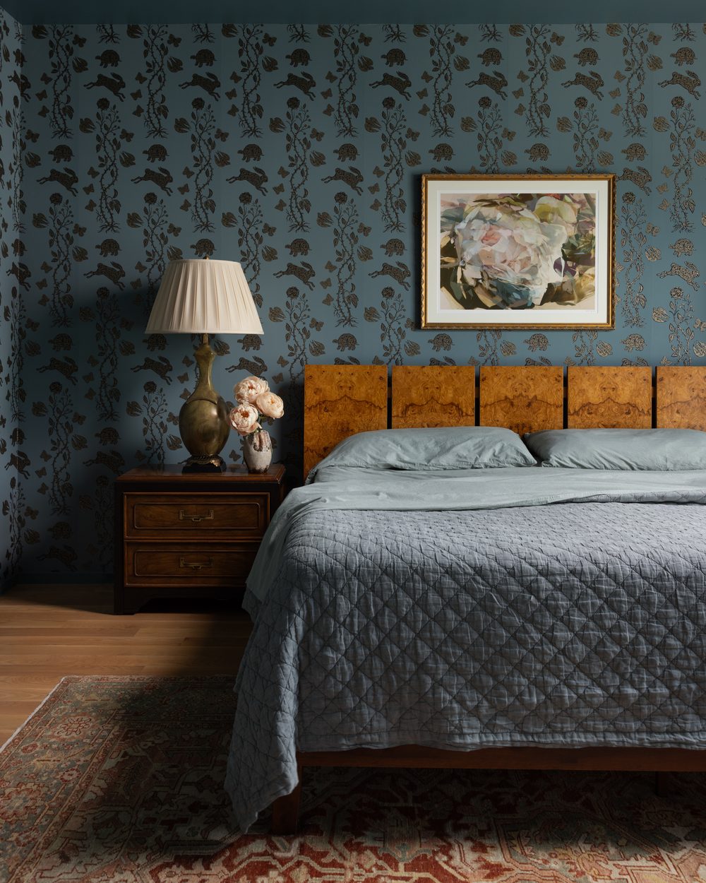 A bedroom with hare-and-turtle wallpaper on slate blue walls, a burl wood headboard, a vintage lamp, and a vintage rug
