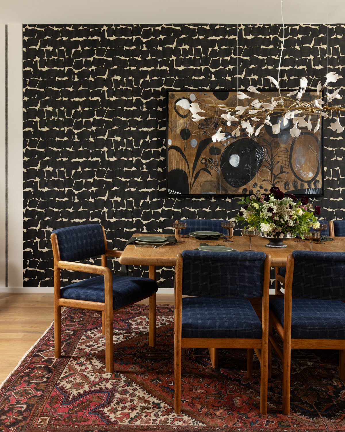 The dining room: bold black brushstroke wallpaper behind an oak table with navy plaid chairs, an abstract painting, a sculptural porcelain chandelier, and a red vintage rug