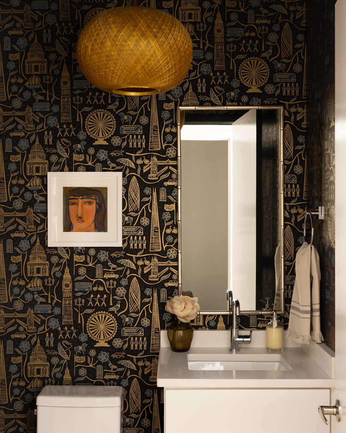A powder room with patterned black-and-gold wallpaper, a woven rattan pendant, and framed vintage art