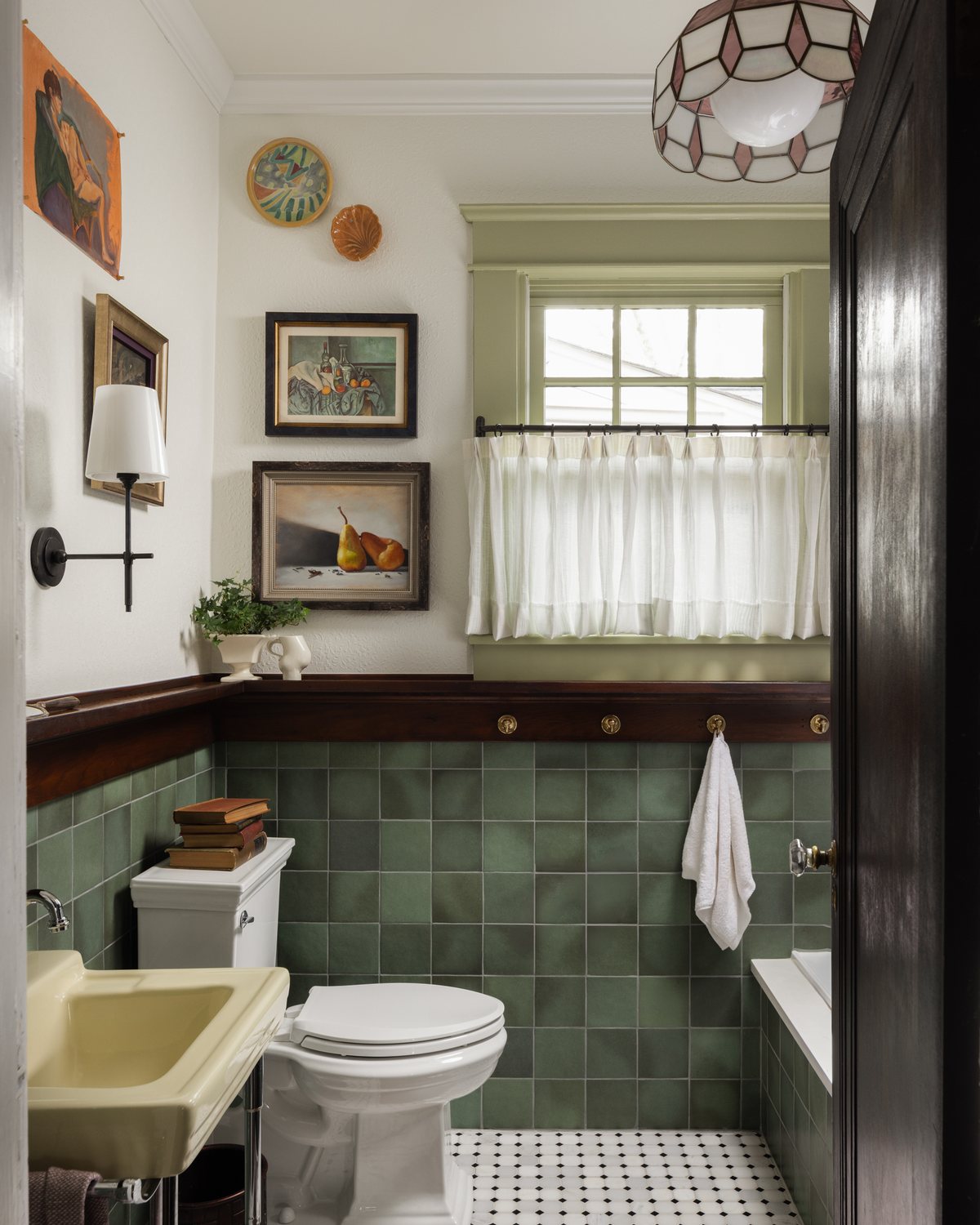 A bathroom with green square tile, a vintage yellow sink, a stained-glass pendant, and collected art