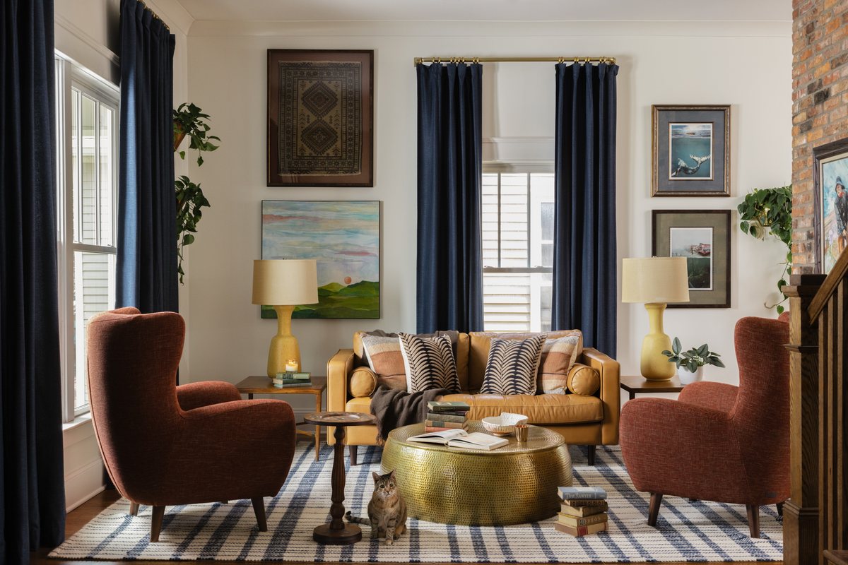 A living room with a camel leather sofa, rust wingback chairs, a hammered brass coffee table, and a plaid rug