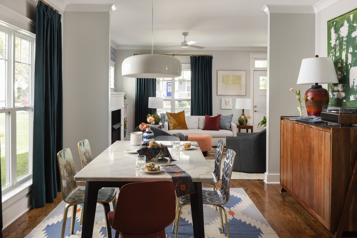 An open concept dining and living space with a marble table, painted plywood chairs, and teal drapes